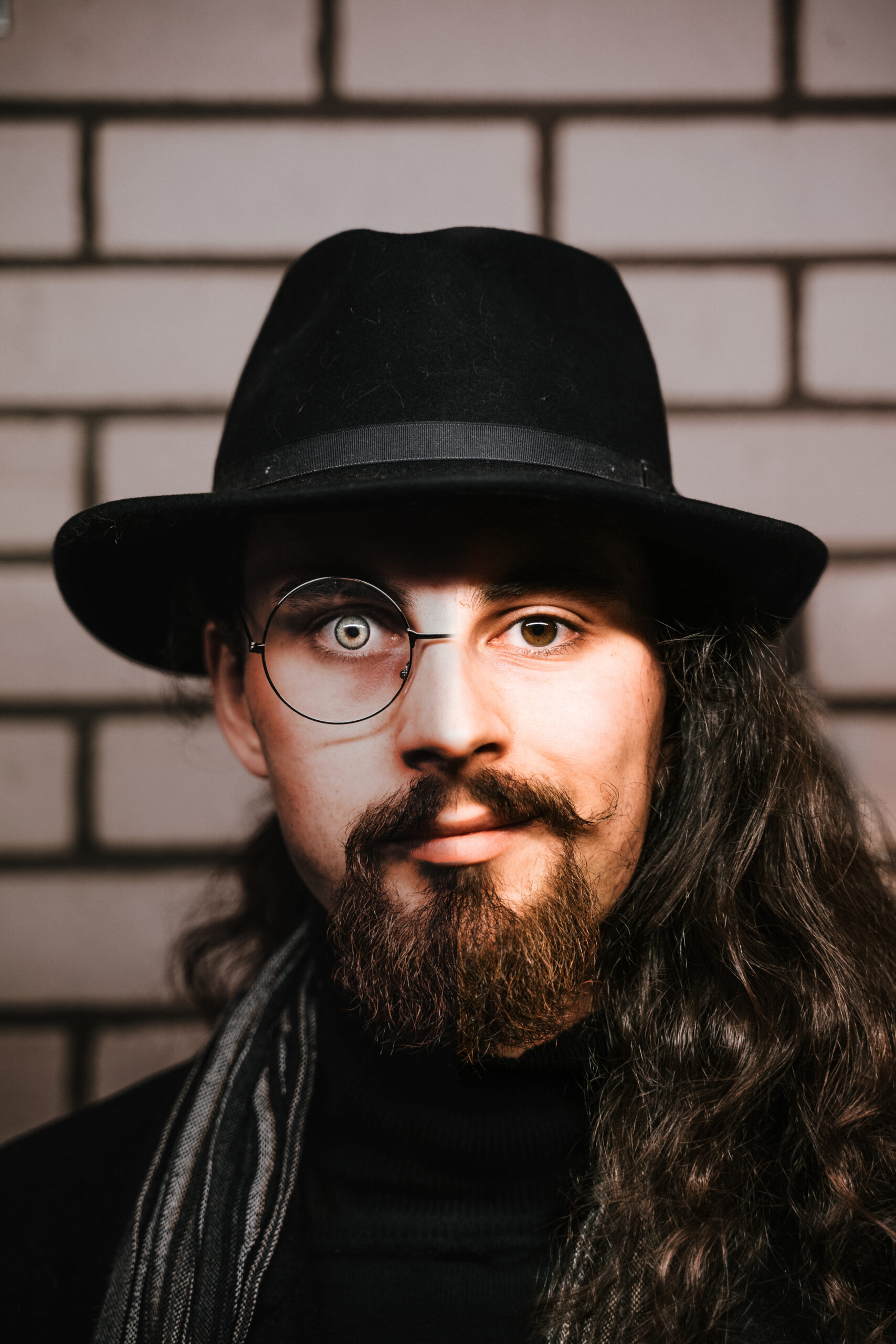 Foto eines Mannes mit einem schwarzen Hut. Auf der rechten Gesichtshälfte trägt er eine (halbe) Brille, das Auge ist sehr hell und die Haare sind zurückgekämmt. Auf der linken Gesichtsseite ist keine Brille zu sehen, die Augenfarbe ist dunkel und die Haare fallen über die Schulter.