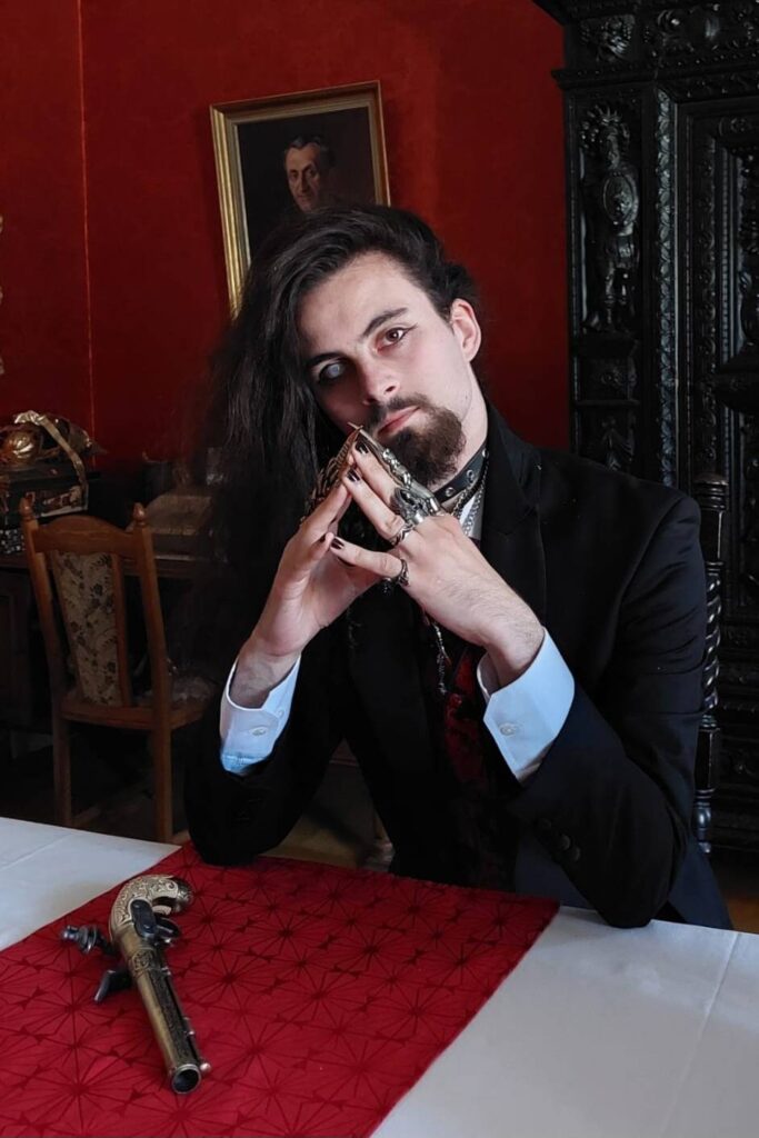 Foto eines Mannes, der die Finger vor der Brust aneinander gelegt hat. Er hat lange, dunkle Haare, trägt eine schwarze Jacke und darunter ein weißes Hemd, außerdem mehrere Ringe