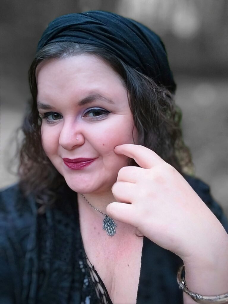 Foto einer Frau, die ein berüschtes, schwarzes Oberteil und ein schwarzes Kopftuch trägt. Um den Hals trägt sie eine Kette mit einem Schutzsymbol. Sie blickt verschmitzt in die Kamera.