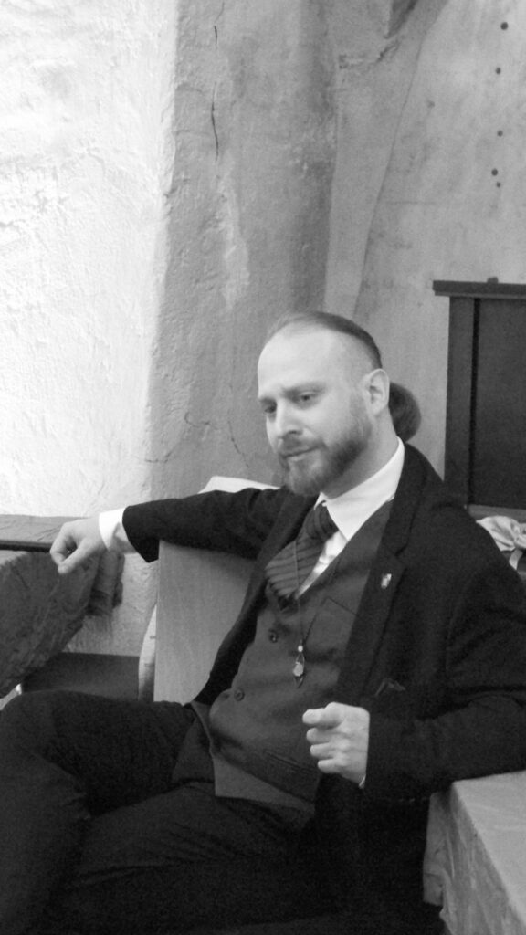 Foto eines Mannes in schwarz/weiß, er trägt einen dreiteilligen Anzug, das lange Haar zurückgebunden, dazu Bart und blickt skeptisch zur Seite