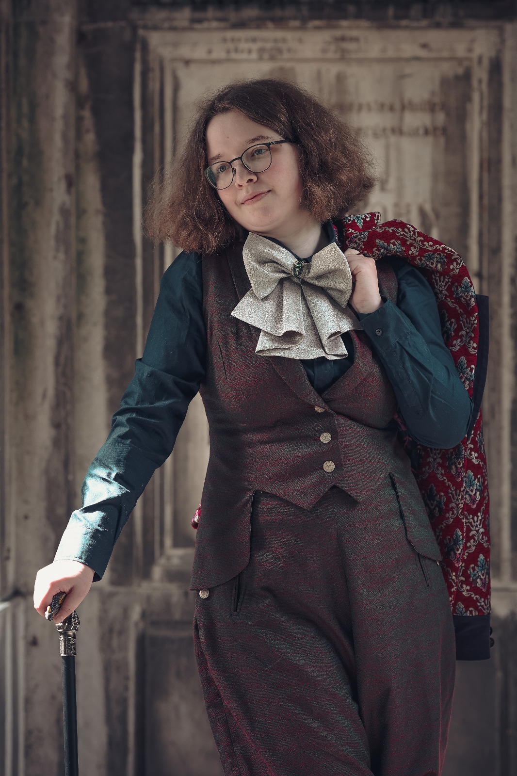 Das Bild zeigt eine Frau mit lockigem, mittellangem Haar, die eine dunkelrote Weste und Hose trägt. Sie trägt eine große, cremefarbene Schleife und eine Brille. Über ihrem Arm hängt ein rot-gold bestickter Mantel. In der Hand hält sie einen Spazierstock. Der Hintergrund ist eine verwitterte Tür.